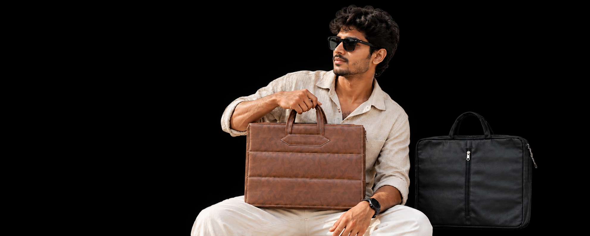 Model in cream clothing holding a brown patterned leather office bag, with a black laptop bag beside him on a black background